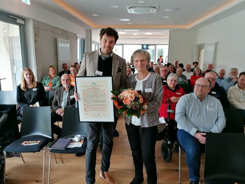 Ein Mann im Anzug überreicht einer Frau in einem geblümten Kleid eine Urkunde, umgeben von einem sitzenden Publikum in einem Konferenzraum. Die Frau hält einen Strauß Blumen in der Hand.