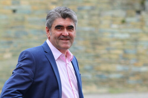 Ein Mann in einer blauen Anzugsjacke und einem rosa Hemd lächelt vor einer Steinmauer im Hintergrund.