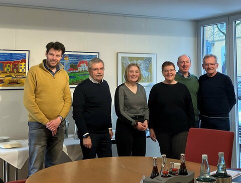 Sechs Personen versammeln sich hinter einem Holztisch in einem gemütlichen Raum, in dem drei bunte Gemälde im Hintergrund zu sehen sind.