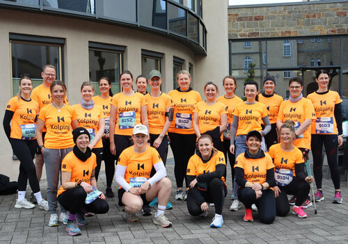 Eine Gruppe von Kolleg*innen von 15 Personen in orangefarbenen "Kolping"-Shirts und schwarzen Shorts posiert für ein Foto vor dem Hotel Aspethera.