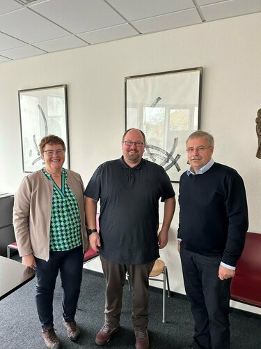 Drei Personen lächeln in einem modernen Büro mit weißen Wänden, schwarz-weißem Kunstwerk und vor einem roten Stühlen.