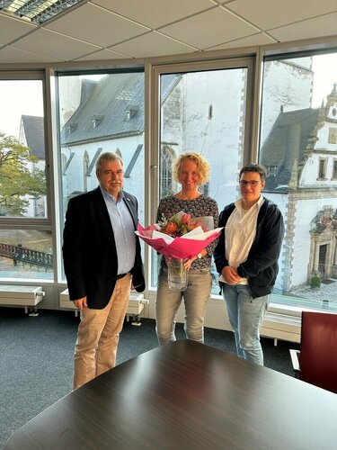 Drei Personen, Hauptgeschäftsführung, Geschäftsführung der Kita und die Prokuristin dieser, stehen in einem Büro mit großen Fenstern und halten einen Blumenstrauß.