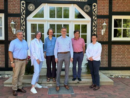 Sechs Personen stehen vor und auf einer Treppe vor einem roten Backsteingebäude mit weißer Tür und dunkelbrauner Verkleidung, im Hintergrund von einer Topfpflanze begleitet.