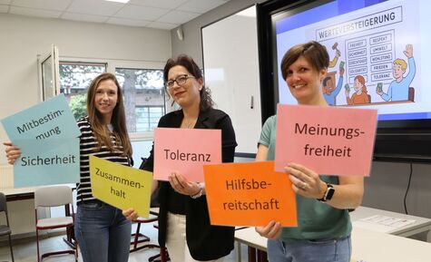 Drei Frauen stehen in einem Klassenzimmer und halten bunte Karten in der Hand, die mit demokratischen Werten beschriftet sind.